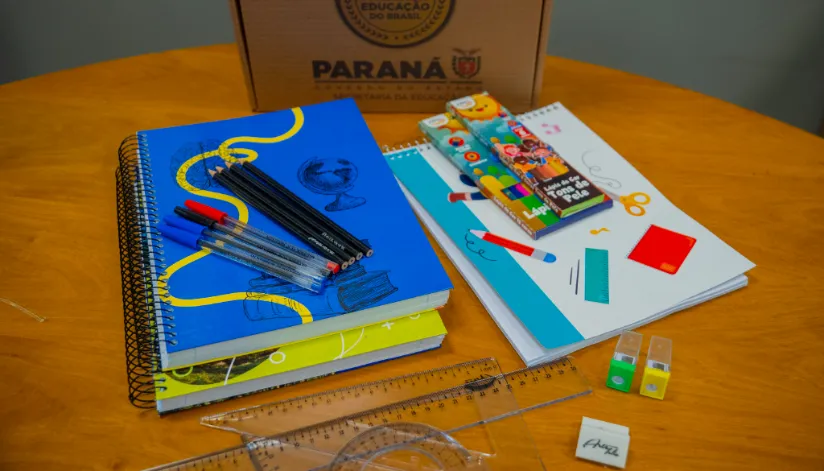 Kit de material escolar sobre uma mesa de madeira, contendo cadernos, canetas, lápis, régua, borrachas e outros itens. Ao fundo, uma caixa de papelão com o logotipo do Governo do Paraná, indicando a entrega de materiais escolares para alunos da rede estadual