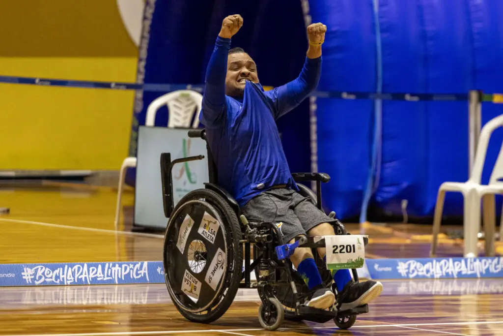 Atleta paralímpico em cadeira de rodas comemorando com os braços erguidos durante uma competição de bocha em um ginásio coberto, com faixa azul ao fundo contendo a hashtag #BocciaParalímpica