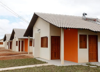 Conjunto de casas populares de um programa habitacional, com telhados inclinados, portas e janelas de madeira, paredes em tons de bege e laranja. Uma das casas possui um aquecedor solar no telhado