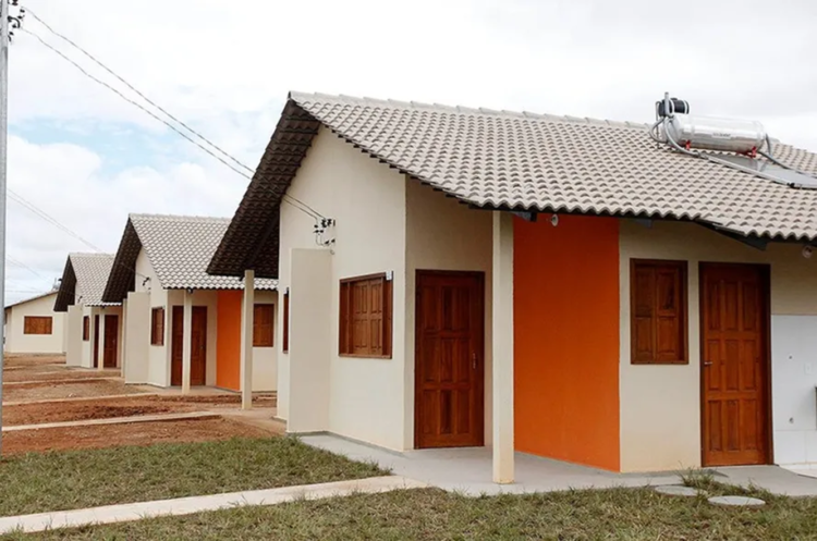 Conjunto de casas populares de um programa habitacional, com telhados inclinados, portas e janelas de madeira, paredes em tons de bege e laranja. Uma das casas possui um aquecedor solar no telhado