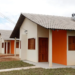 Conjunto de casas populares de um programa habitacional, com telhados inclinados, portas e janelas de madeira, paredes em tons de bege e laranja. Uma das casas possui um aquecedor solar no telhado