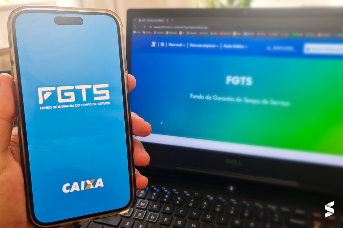 Imagem de celular mostrando o aplicativo do FGTS da Caixa Econômica Federal, com o fundo do site oficial do FGTS visível em um laptop.