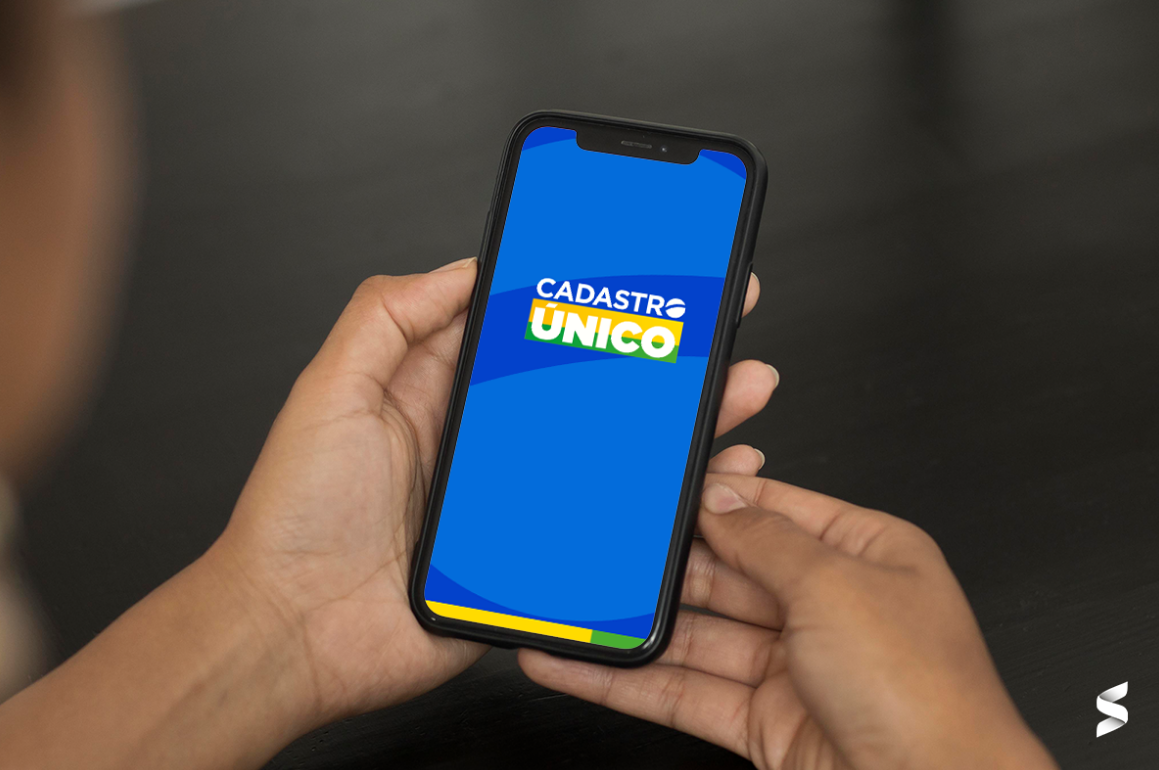 Tela do CadÚnico aberta em um smartphone, com a logo visível ao fundo em cores azul, amarela e verde.