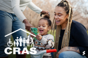 Mãe e filha interagindo com um celular enquanto uma pessoa as apoia, com o símbolo do CRAS na imagem, representando o agendamento de serviços sociais pelo WhatsApp.