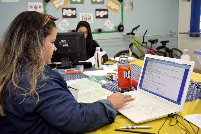 Mulher utilizando um computador para realizar o agendamento no CRAS, com documentos e outros materiais de apoio à mesa.