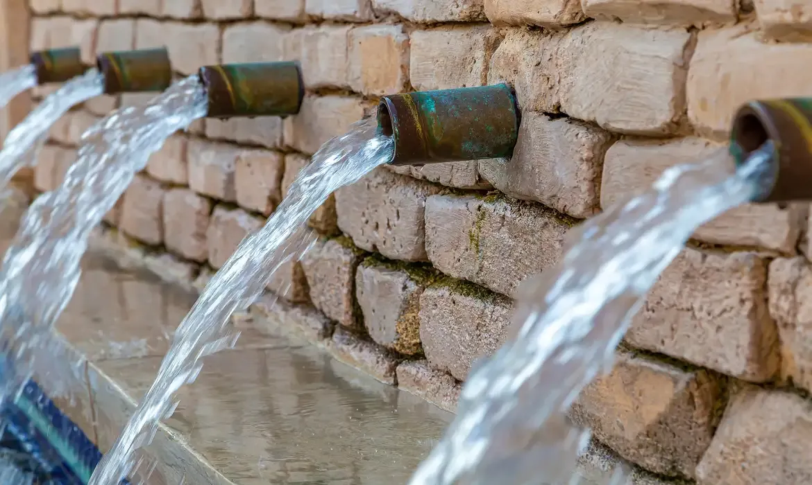 Água saindo de canos metálicos embutidos em uma parede de tijolos.