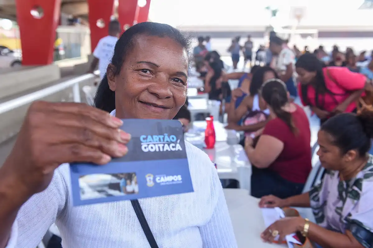  Mulher sorridente segurando o Cartão Goitacá durante evento de distribuição do benefício.