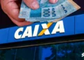 Mão segurando notas de dinheiro em frente à agência da Caixa Econômica Federal