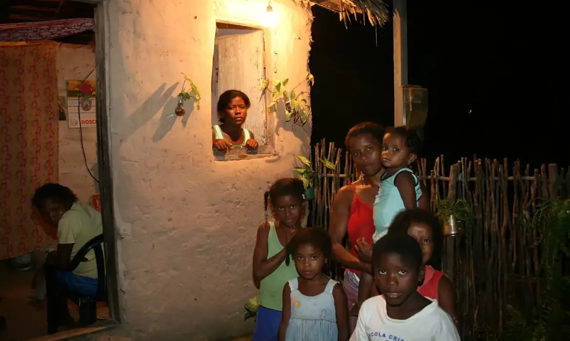 Família de baixa renda em frente a uma casa iluminada à noite.