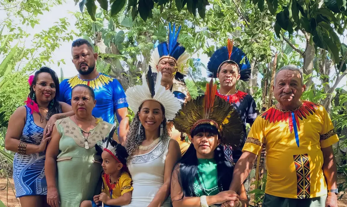 Grupo de pessoas indígenas vestindo trajes tradicionais e adornos de penas, posando em meio à natureza.