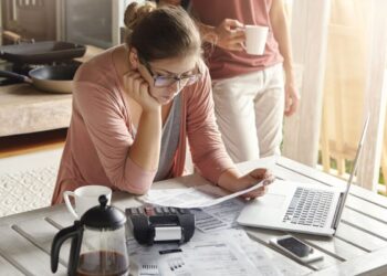 Mulher jovem analisando contas e documentos financeiros enquanto utiliza uma calculadora e um laptop, representando a consulta de dívidas online.