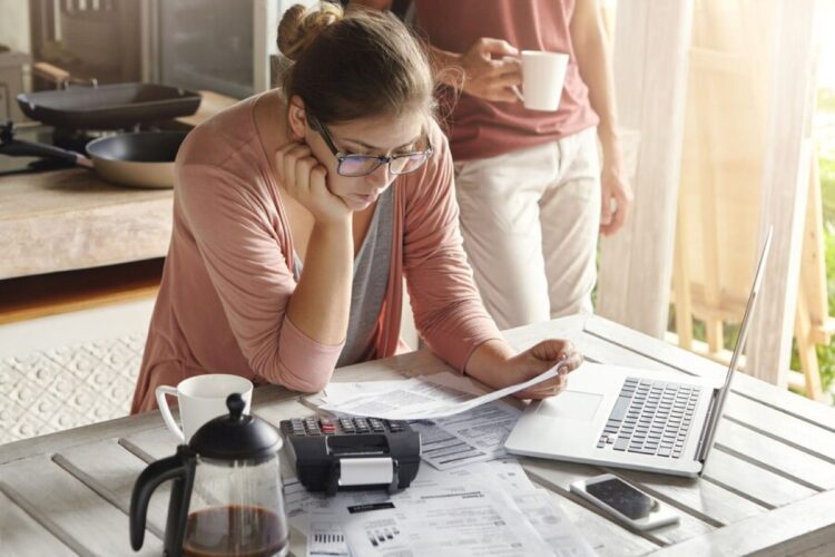 Mulher jovem analisando contas e documentos financeiros enquanto utiliza uma calculadora e um laptop, representando a consulta de dívidas online.