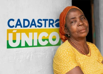 Mulher de pele negra e lenço laranja encostada em uma parede com o logo do Cadastro Único ao fundo