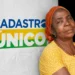 Mulher de pele negra e lenço laranja encostada em uma parede com o logo do Cadastro Único ao fundo