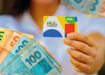 Cartão do Bolsa Família sendo segurado por uma pessoa, com notas de dinheiro ao fundo