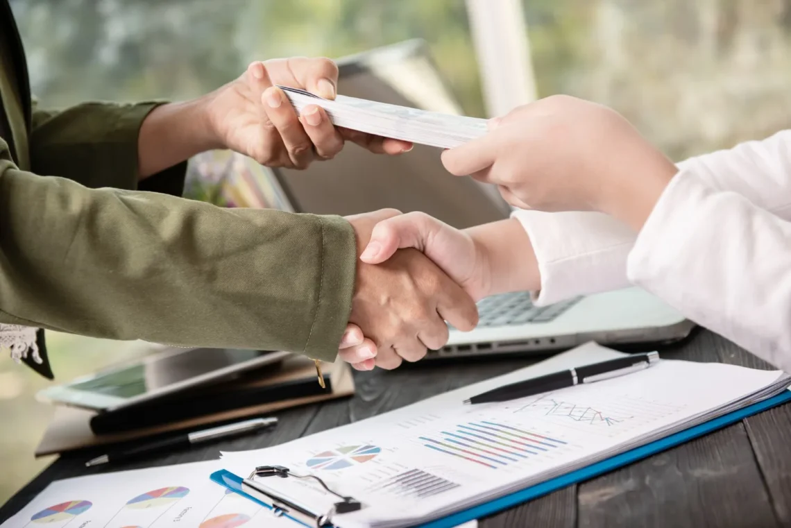 Duas pessoas apertando as mãos em um acordo financeiro, com dinheiro sendo entregue e documentos financeiros sobre a mesa