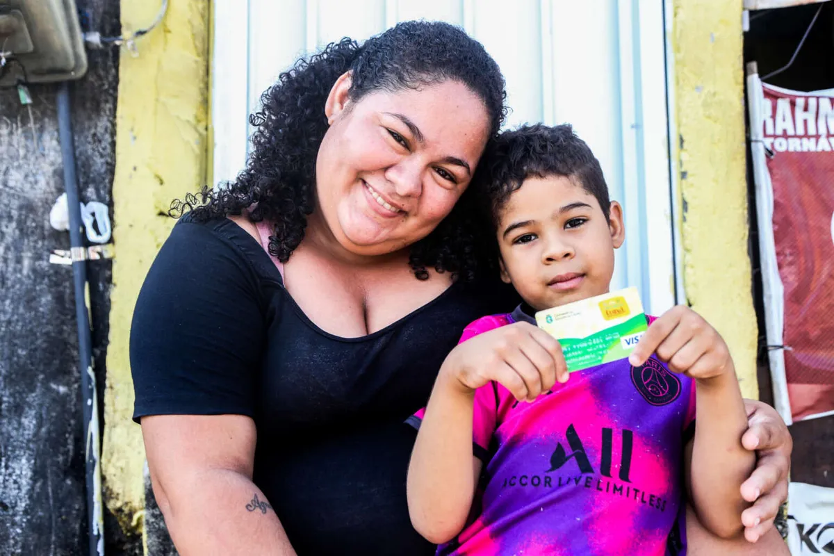  Mulher e criança segurando o Cartão Mais Infância, representando a entrega do benefício às famílias.