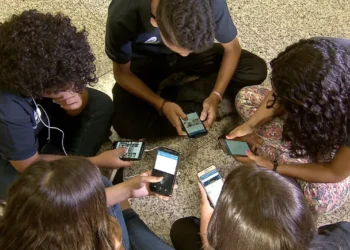 Estudantes usando celulares no ambiente escolar durante o intervalo, representando o debate sobre o uso de dispositivos móveis nas escolas.