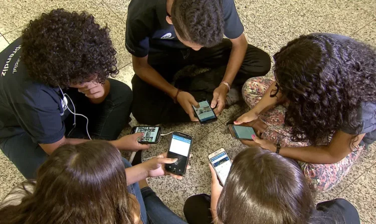 Estudantes usando celulares no ambiente escolar durante o intervalo, representando o debate sobre o uso de dispositivos móveis nas escolas.