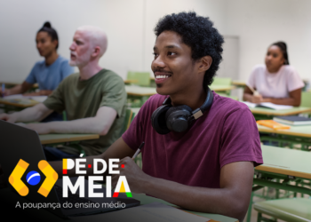 Estudantes em sala de aula com foco no jovem sorrir enquanto usa laptop, representando o pagamento de até R$ 9.200 do programa Pé-de-Meia para estudantes.