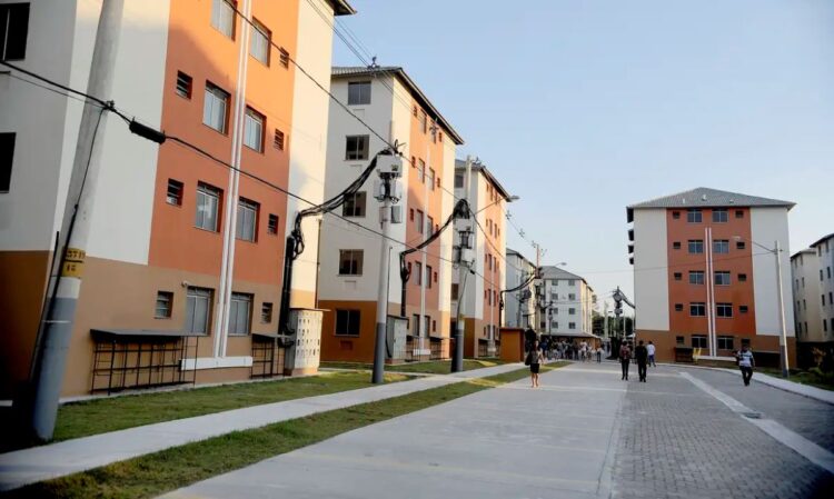 Imagem de conjunto habitacional do programa Minha Casa, Minha Vida, com moradias e pessoas caminhando na área externa.