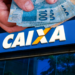 Mãos segurando cédulas de 100 reais na frente de uma agência da CAIXA, símbolo de benefício que começa a ser liberado esta semana.
