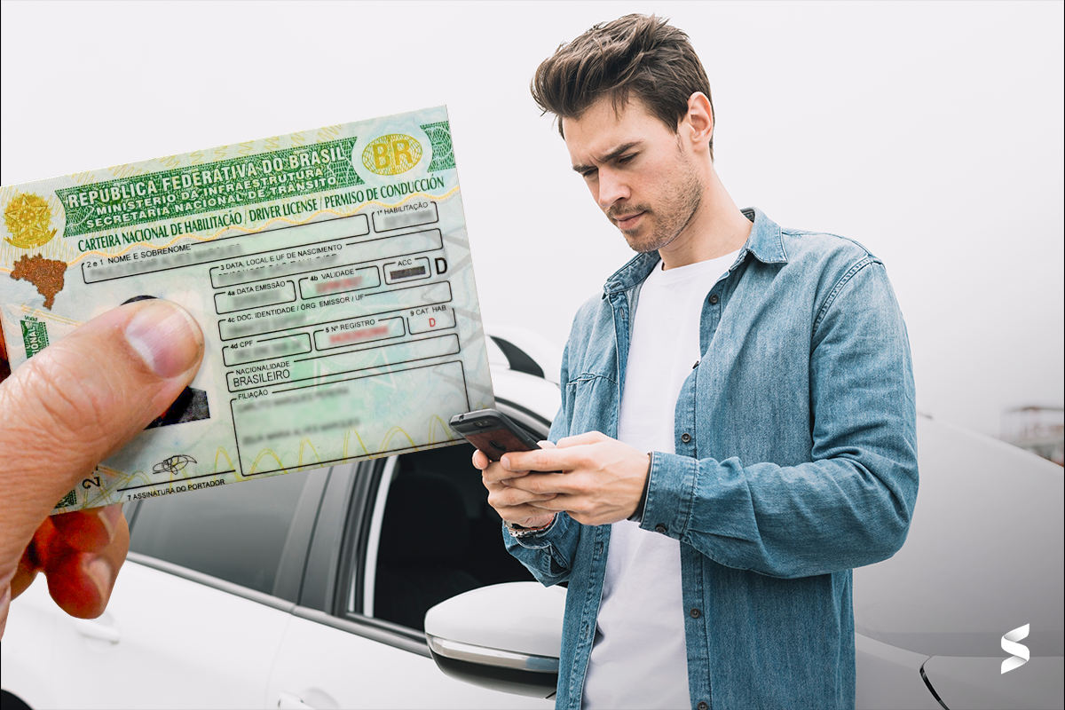 Carteira Nacional de Habilitação brasileira sendo segurada por uma mão em primeiro plano, com um homem ao fundo usando celular próximo a um carro.