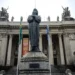 Estátua em frente à Assembleia Legislativa do Rio de Janeiro, com a fachada imponente ao fundo, representando o cenário do feriado de Tiradentes.