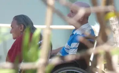 "Casal de idosos, um em cadeira de rodas, em ambiente tranquilo, sugerindo cuidado e apoio mútuo.