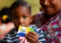 "Criança segurando o cartão do Programa Bolsa Família, com a mãe ao fundo."