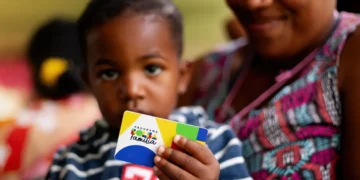 "Criança segurando o cartão do Programa Bolsa Família, com a mãe ao fundo."