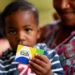 "Criança segurando o cartão do Programa Bolsa Família, com a mãe ao fundo."