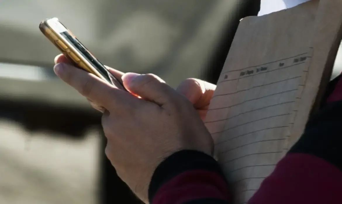 Pessoa segurando um celular e um bloco de anotações ao ar livre.