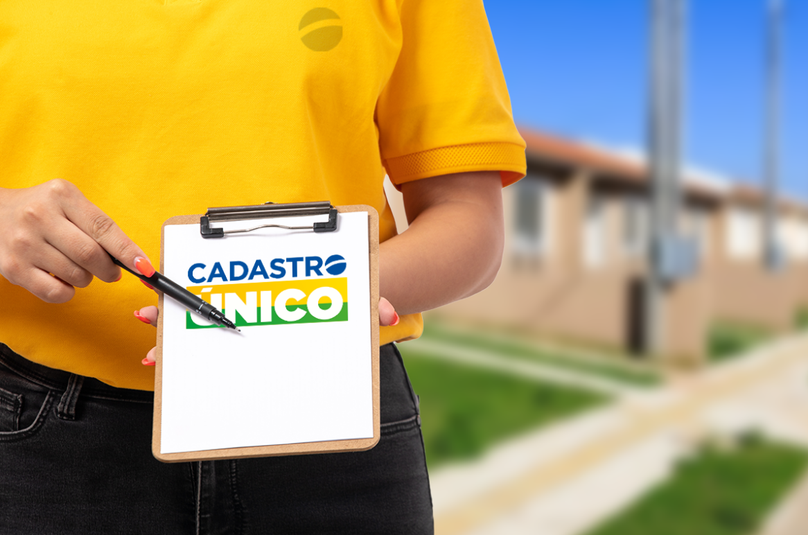 Pessoa com camiseta amarela segurando uma prancheta com o logo do CadÚnico, com casas ao fundo.