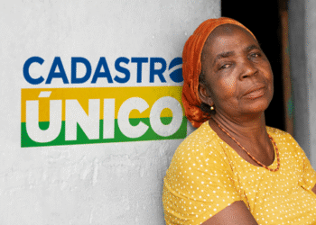 Mulher idosa em frente a parede com logotipo do Cadastro Único, representando beneficiários dos programas sociais em 2025.