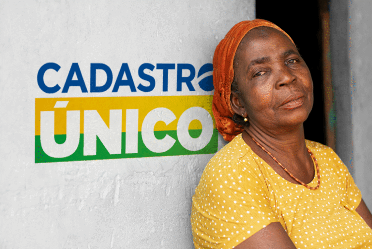Mulher idosa em frente a parede com logotipo do Cadastro Único, representando beneficiários dos programas sociais em 2025.