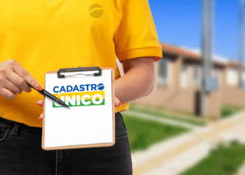 Pessoa segurando uma prancheta com o logo do CadÚnico em frente a um conjunto habitacional.