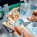 Pessoa retirando dinheiro de um caixa eletrônico, com cédulas de reais em destaque, representando o tema sobre o CPF no Pé-de-Meia