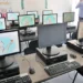 Sala de informática com computadores recém-instalados em funcionamento, exceto um monitor desligado.