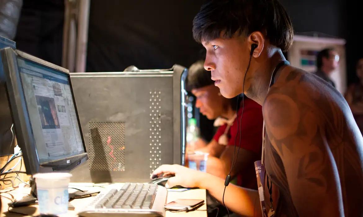 Jovem indígena utilizando computador em ambiente coletivo, com fone de ouvido e atenção à tela.