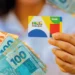 Pessoa segurando cartão do Bolsa Família com notas de real ao fundo.