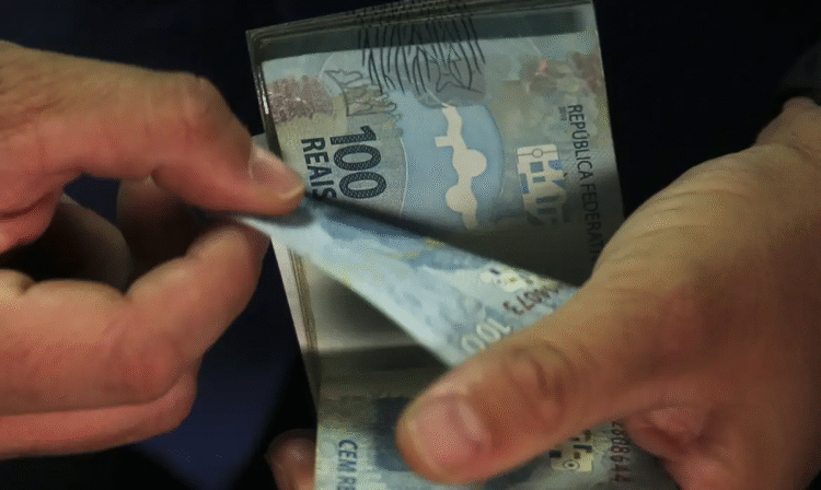 Pessoa segurando cédulas de R$ 100, simbolizando o valor do décimo terceiro salário de um salário mínimo.