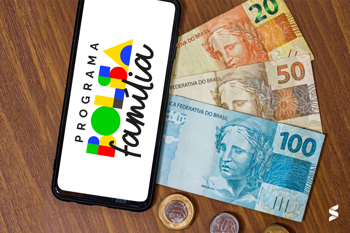Imagem de celular mostrando o logo do Bolsa Família, com notas de dinheiro e moedas ao fundo.