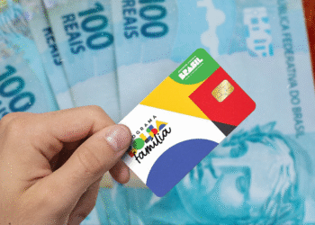 Imagem de um cartão do Bolsa Família sendo segurado por uma mão, com notas de R$ 100 ao fundo.