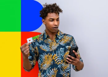 Homem segurando o cartão do Bolsa Família e olhando para o celular em frente a um fundo colorido.