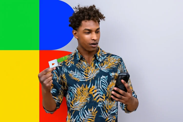 Homem segurando o cartão do Bolsa Família e olhando para o celular em frente a um fundo colorido.