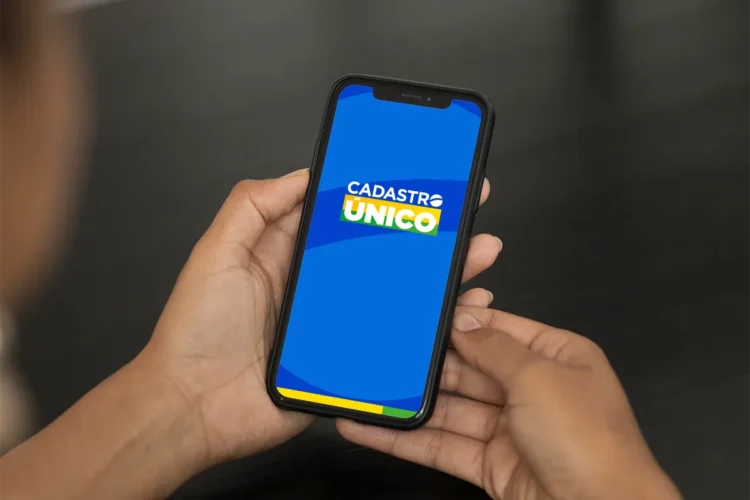 Pessoa segurando celular com tela do aplicativo CadÚnico aberto, em fundo azul com logo e cores do Brasil.