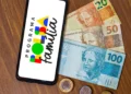 Celular exibindo o logo do Programa Bolsa Família, com notas de 20, 50 e 100 reais ao fundo