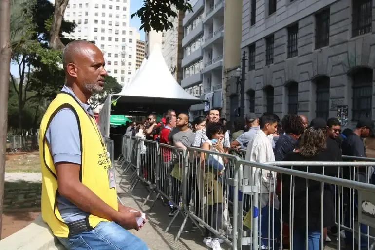 "Fila de pessoas aguardando atendimento em evento de mutirão, com atendente de colete amarelo organizando a fila."