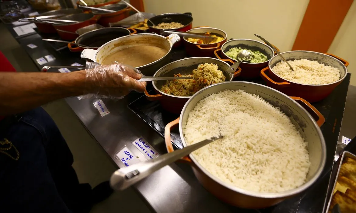 Servidor manipula alimentos em refeitório popular com várias opções de comida
